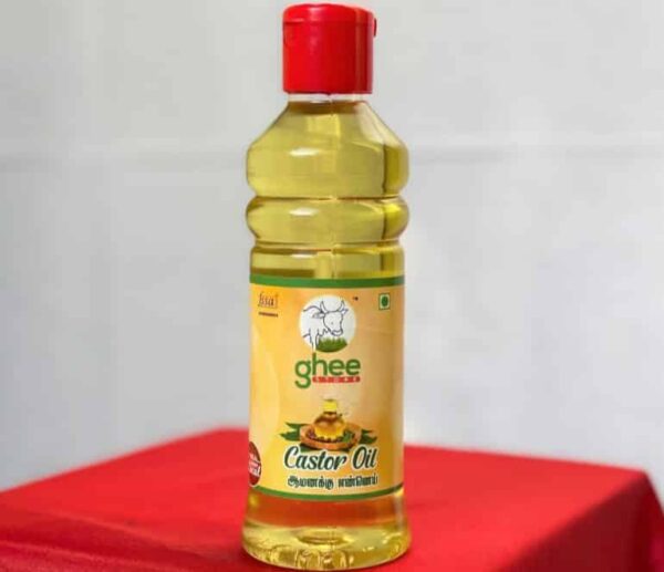 A bottle of castor oil with a red cap, featuring a label that reads "ghee" and "Castor Oil" in Tamil, placed on a red surface.
