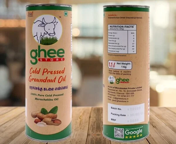 A cylindrical container of cold pressed groundnut oil, labeled "ghee STORE," featuring a cow illustration and text in both English and Tamil. The container includes nutritional facts on one side, with details on the product's weight (1 kg) and packaging information. It showcases a natural, earthy design with peanuts depicted at the bottom.
