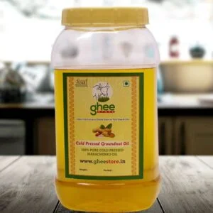 A transparent plastic jar of cold-pressed groundnut oil with a yellow lid and a green label, placed on a wooden surface in a kitchen setting. The label includes the product name, details about purity, and a website.