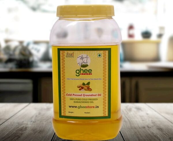 A transparent plastic jar of cold-pressed groundnut oil with a yellow lid and a green label, placed on a wooden surface in a kitchen setting. The label includes the product name, details about purity, and a website.