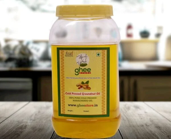 A transparent plastic jar of cold-pressed groundnut oil with a yellow lid and a green label, placed on a wooden surface in a kitchen setting. The label includes the product name, details about purity, and a website.