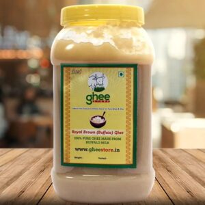 A transparent jar of Royal Brown (Buffalo) Ghee with a yellow lid, featuring a label that reads "ghee store" and indicates it is 100% pure ghee made from buffalo milk. The background is a blurred interior of an establishment.