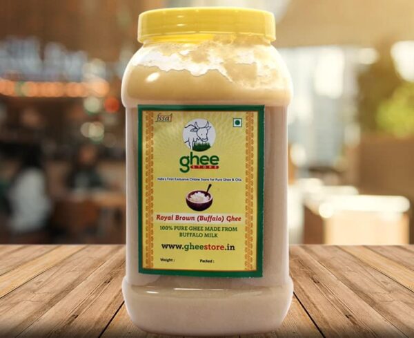 A transparent jar of Royal Brown (Buffalo) Ghee with a yellow lid, featuring a label that reads "ghee store" and indicates it is 100% pure ghee made from buffalo milk. The background is a blurred interior of an establishment.