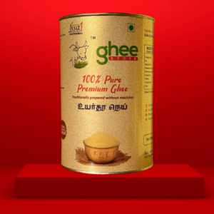 A cylindrical tin of 100% Pure Premium Ghee, featuring a gold and cream label with a cow logo and text in English and Tamil, placed on a red background.