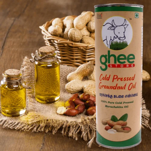 A bottle of cold pressed groundnut oil and two smaller jars of oil, placed next to a woven basket filled with peanuts, all arranged on a textured fabric background.